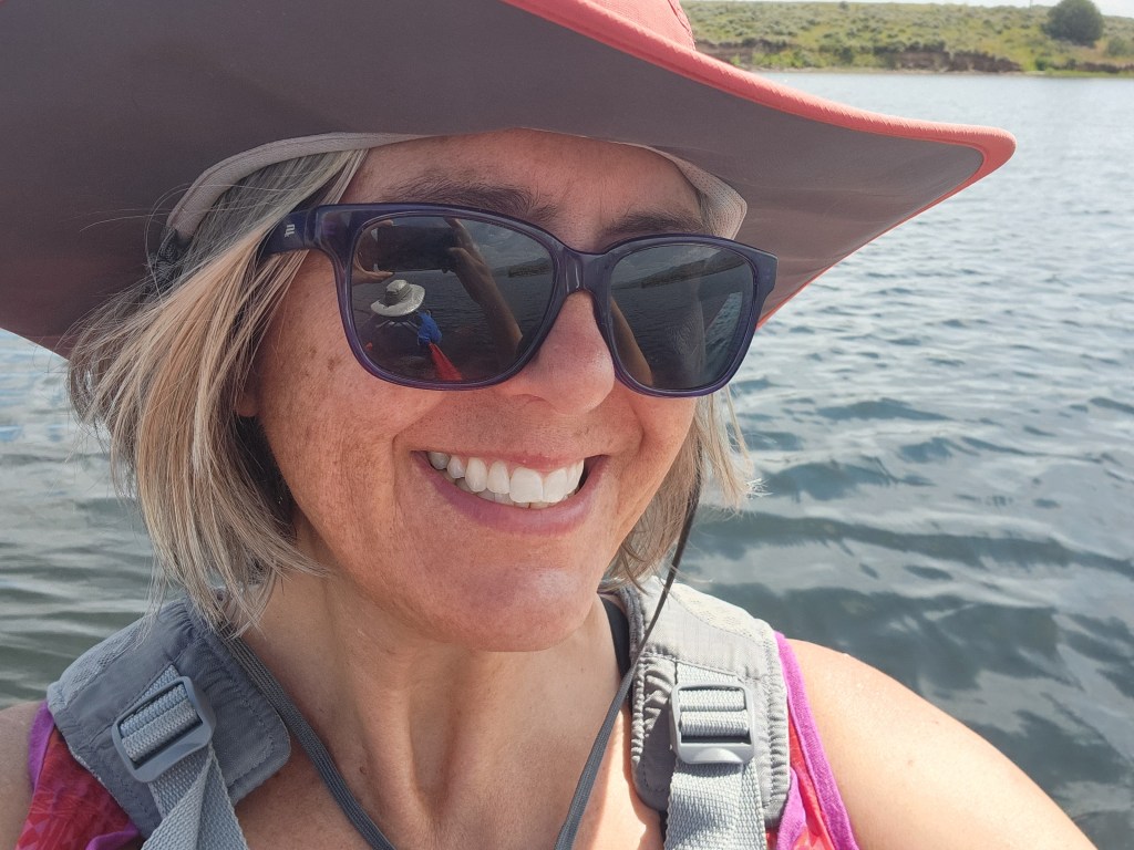 A smiling woman with short, light hair wearing sunglasses and a wide-brimmed hat, standing near a body of water.
